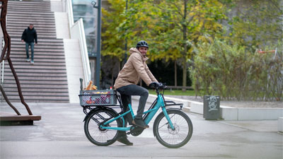 Je ziet een man op een blauwe Cube Longtail Hybride bakfiets door het stadscentrum rijden en voedsel opslaan.