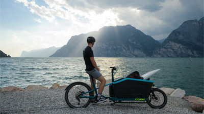 Een man staat met zijn bakfiets op de oever van het meer. Hij kijkt naar de bergen op de achtergrond.