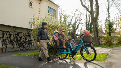 Je ziet een moeder naar haar blauwe bakfiets lopen. Twee kinderen van klanten zitten op de bagagedrager; de situatie is in een woonwijk.