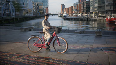 Je ziet een vrouw op een roze Cube Ella Hybrid e-bike. Ze fietst langs een rivier. Er is een stad op de achtergrond.