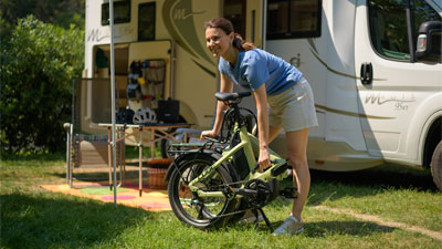 Je ziet een vrouw voor een camper staan. Voor haar staat een opgevouwen groene vouwfiets.