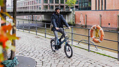 Een man rijdt op zijn blauwe vouwfiets, met huizen en een rivier op de achtergrond.