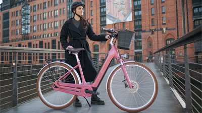 Je ziet een vrouw achter een roze Cube Ella Hybrid e-bike. Er staat een gebouw op de achtergrond.