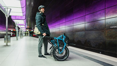 Je ziet een man in een metrostation met een opgevouwen blauwe vouwfiets voor zich.