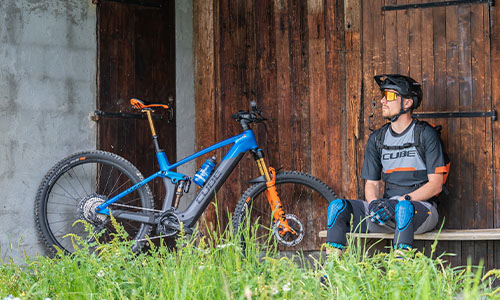 Je ziet een fietser op een bankje zitten voor een schuur. Naast hem staat zijn volledig elektrische mountainbike.