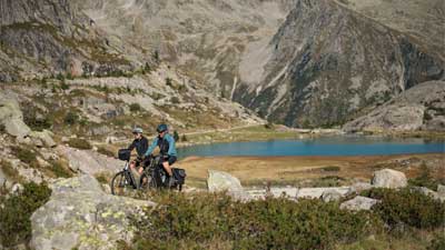 Een stel rijdt op een trekkingfiets door een rotsachtig landschap, met bergen en een meer op de achtergrond.