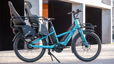 Te zien is een blauwe Cube Longtail Hybride bakfiets met twee uitgeruste kinderzitjes. 