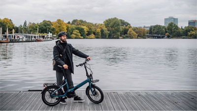 Een man staat naast zijn blauwe vouwfiets op een steiger. Op de achtergrond zijn water en een stad te zien.