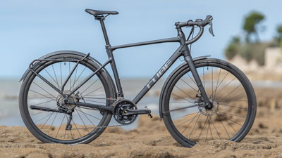 Je ziet een zwarte Cube gravel bike op een rotsachtig landschap staan met de kust op de achtergrond.