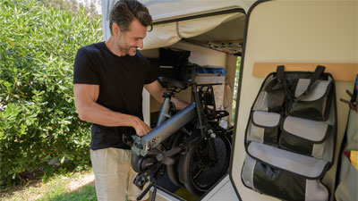 Een man plaatst zijn opgevouwen grijze vouwfiets in zijn open camper.