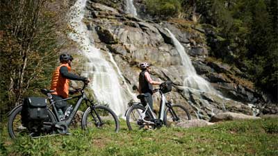 Een stel duwt hun fietsen door een weiland, met een waterval op de achtergrond.