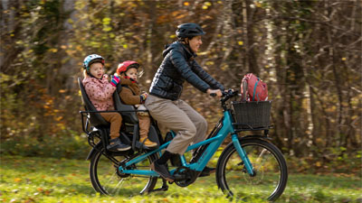 Je ziet een moeder op een blauwe Cube Longtail bakfiets. Twee kleine kinderen zitten op de bagagedrager.